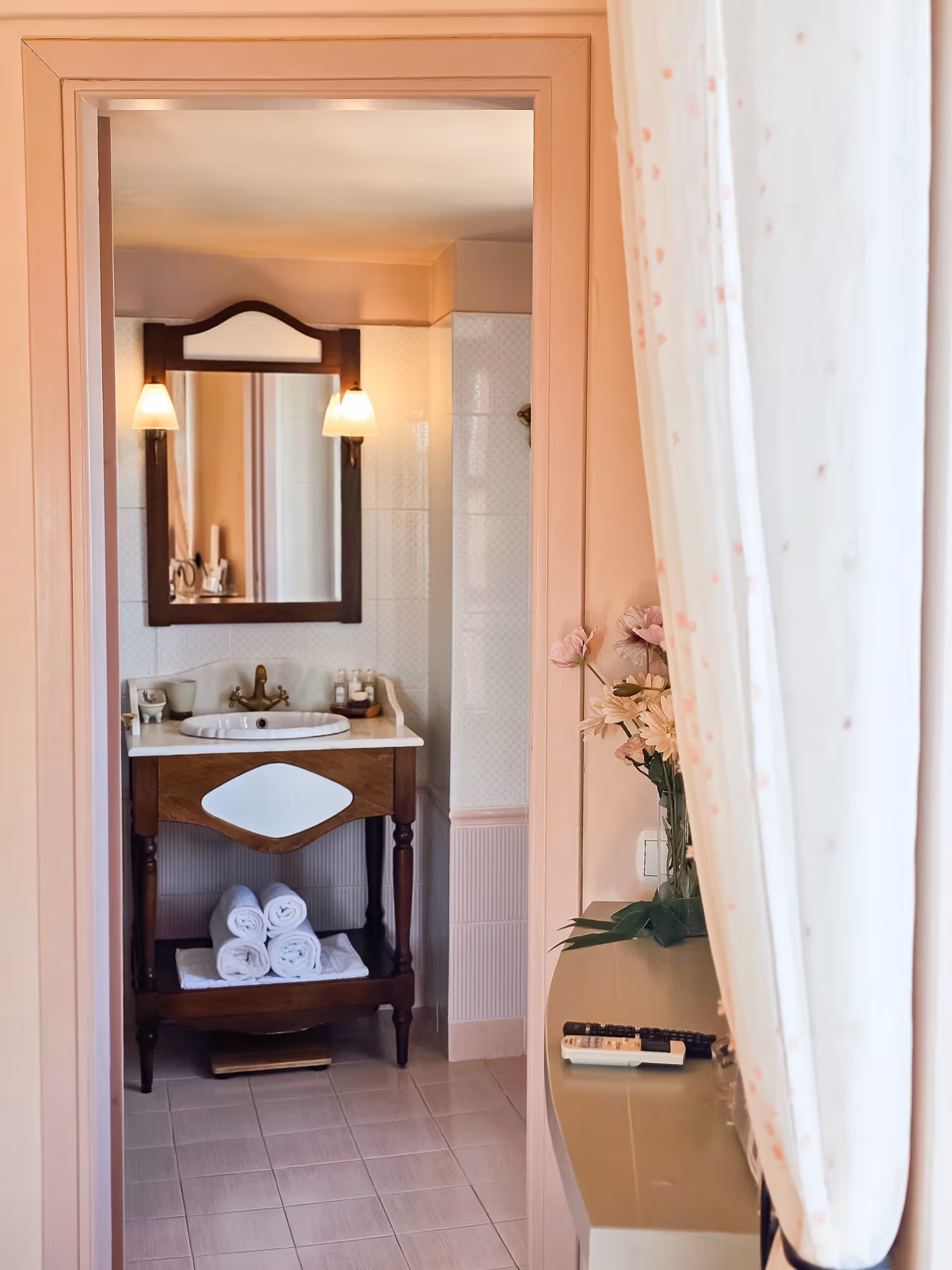 View of a bathroom with wooden sink vanity, mirror, wall lights, towels, and flowers on side table.