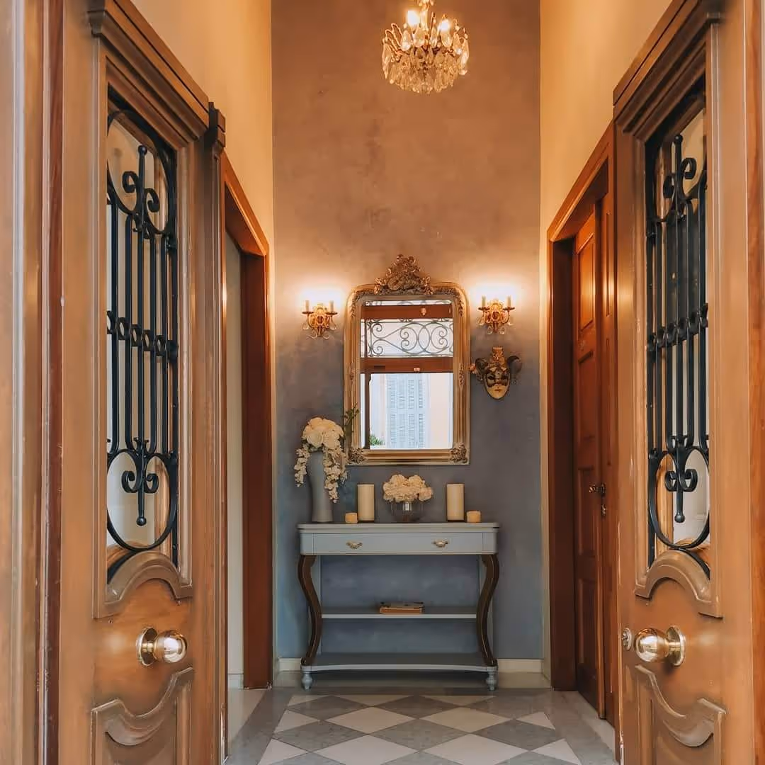 Hallway with a gray console table, a decorative mirror, wall sconces, flowers, and wooden doors partially open.