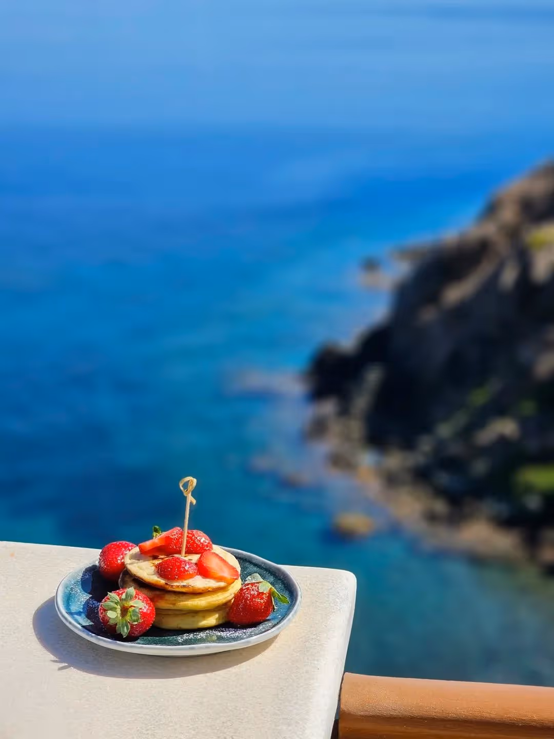 Plate of pancakes topped with strawberries on a ledge with ocean and rocky coastline background.
