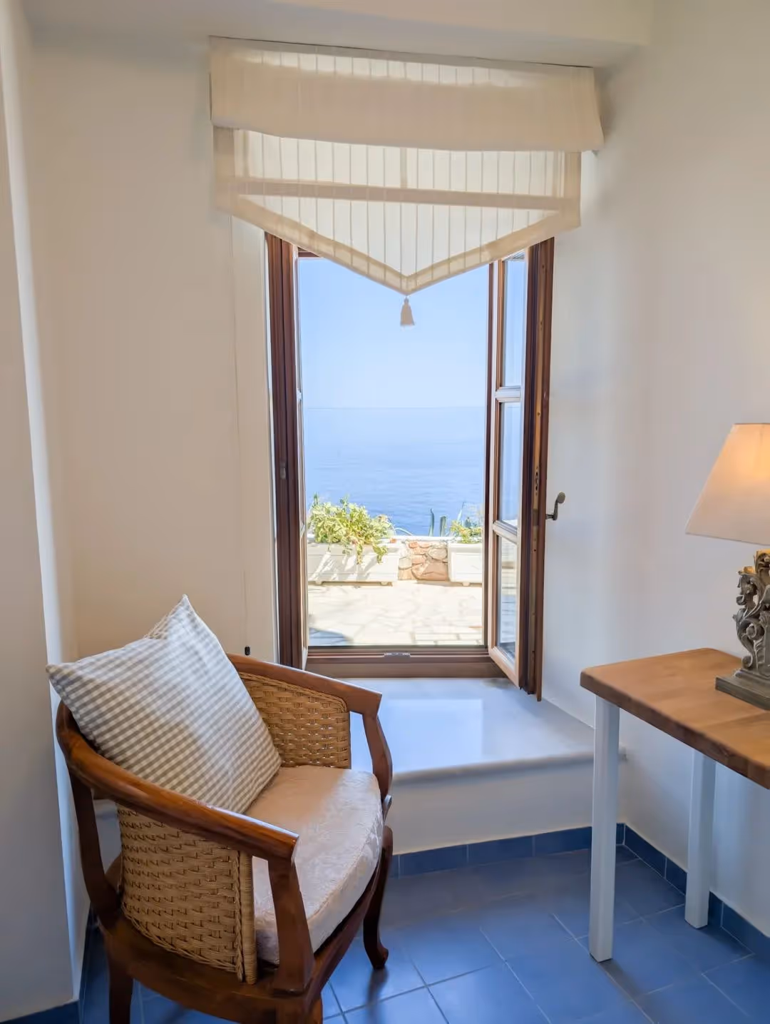 Wooden chair with cushion beside a window showing a sea view and potted plants outside.