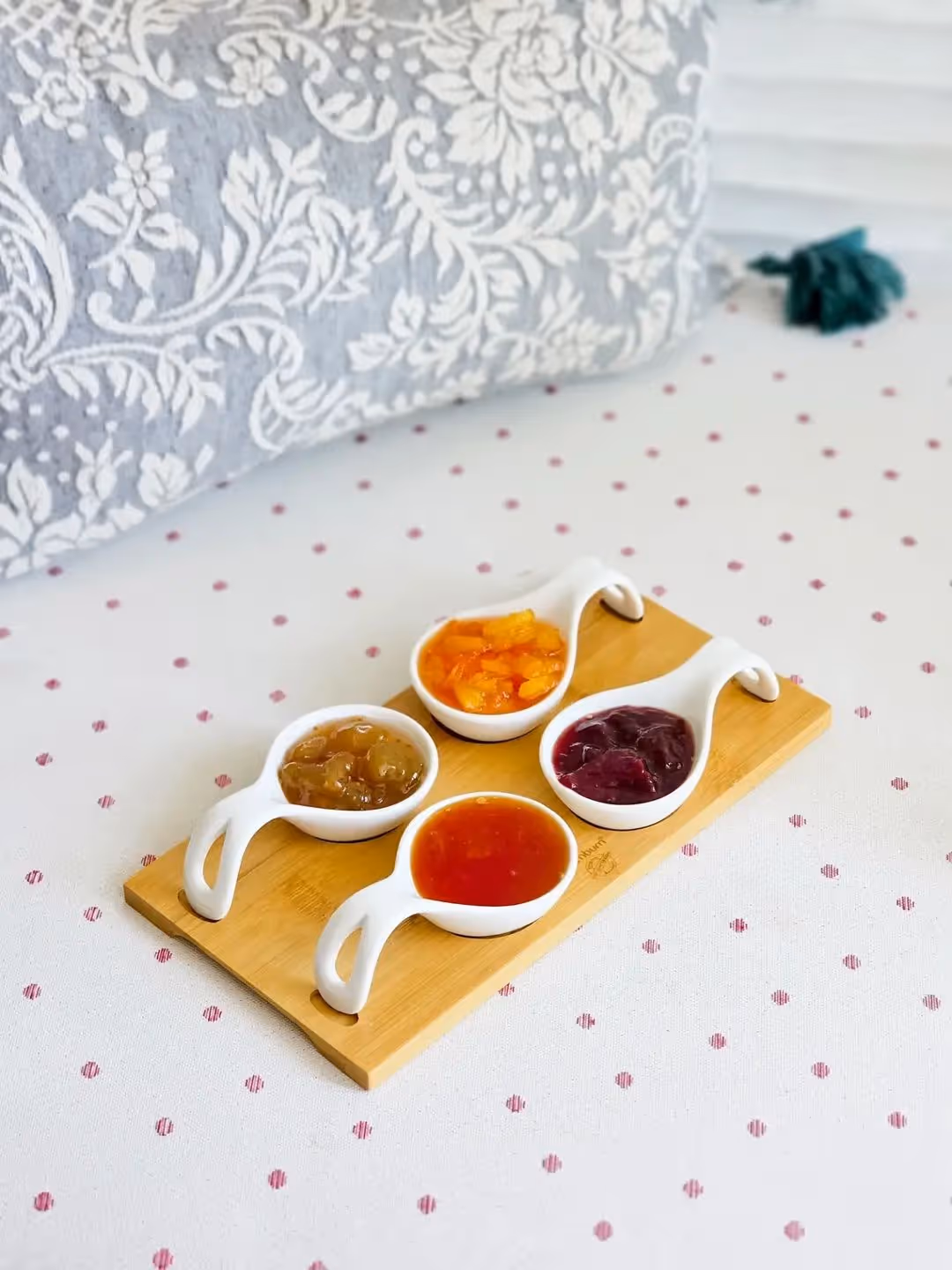 Four white ceramic spoons with different jams on a wooden board on a polka dot tablecloth.