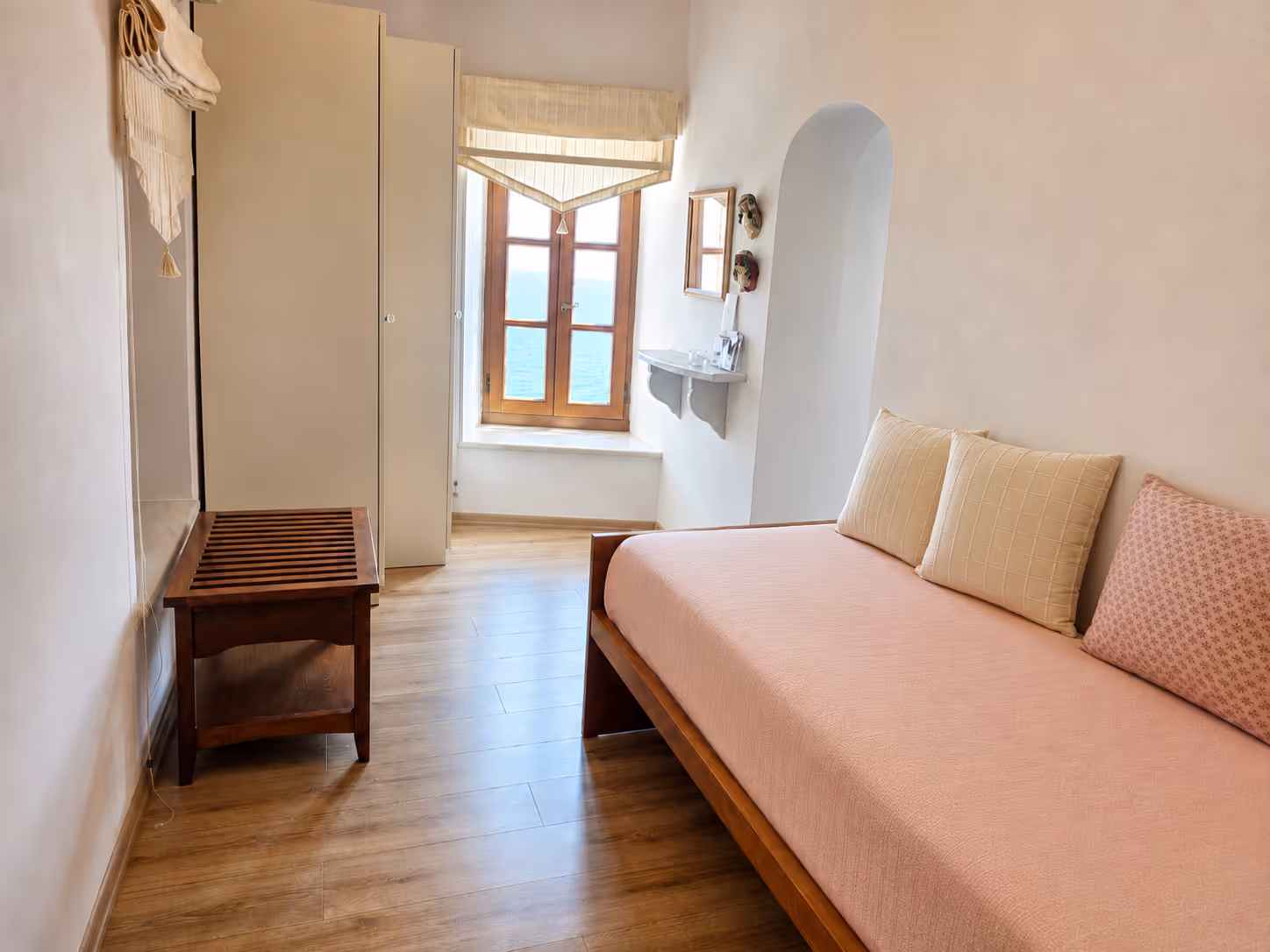 Simple bedroom with wooden floor, daybed with pillows, wardrobe, and window with sea view.