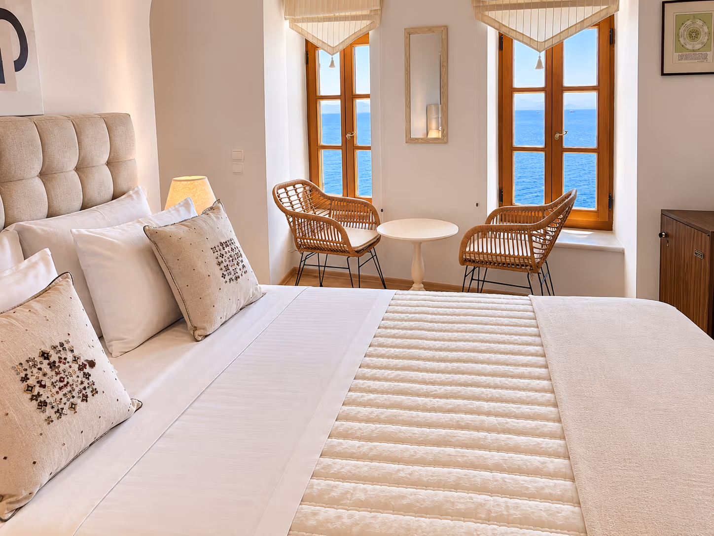 Cozy bedroom with a large bed, beige cushions, two wicker chairs, and windows overlooking the sea.