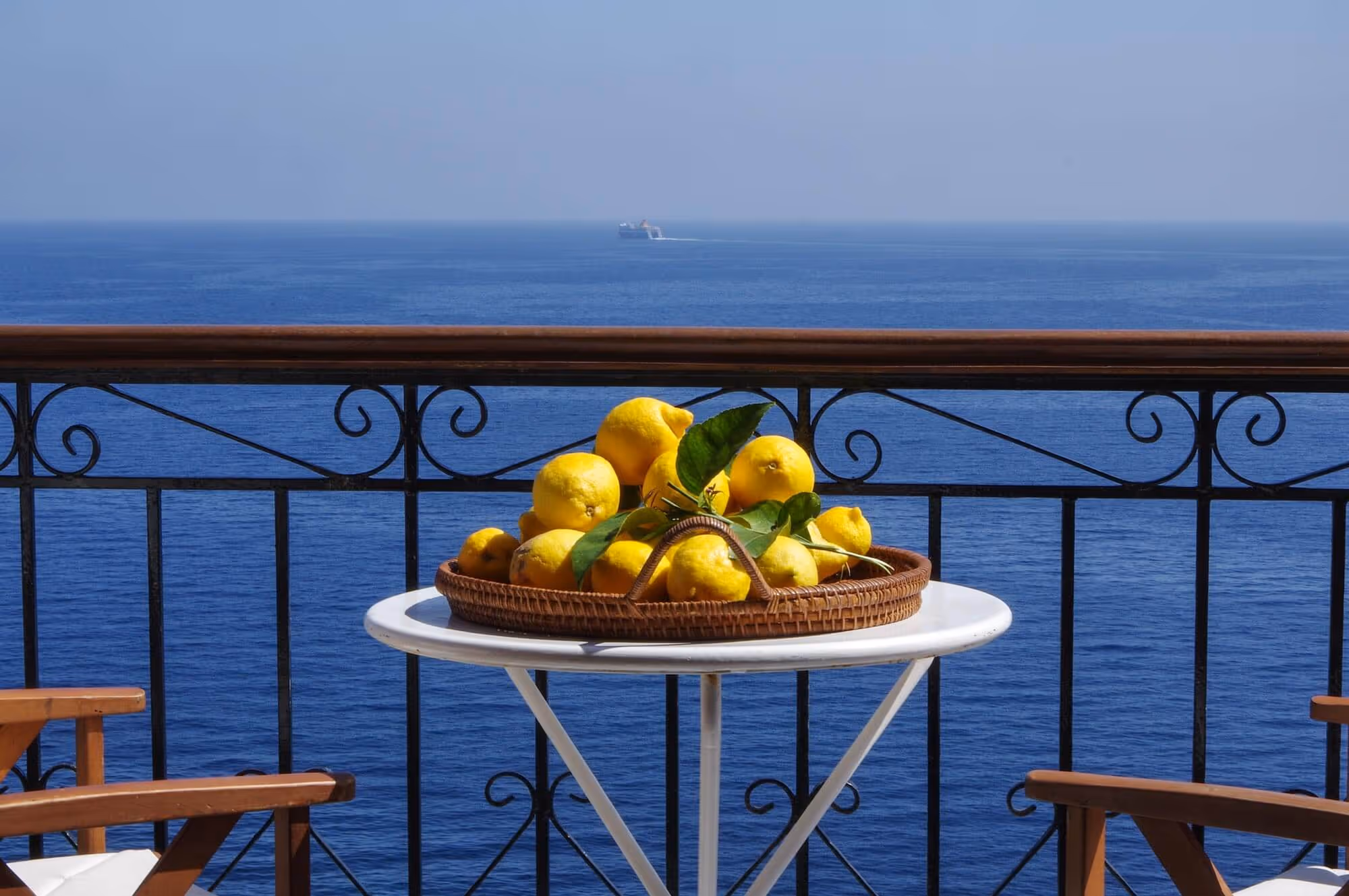 Tray of lemons on a white table on a balcony overlooking a calm blue sea with a distant ship.