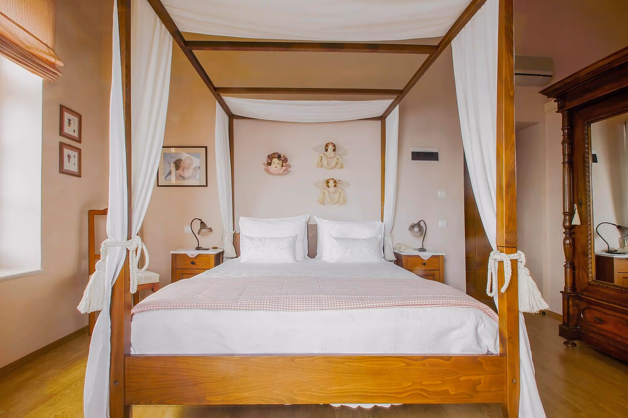 Bedroom with wooden four-poster bed draped with white curtains, two pillows, nightstands with lamps, and wall decorations including framed pictures and angel figurines.