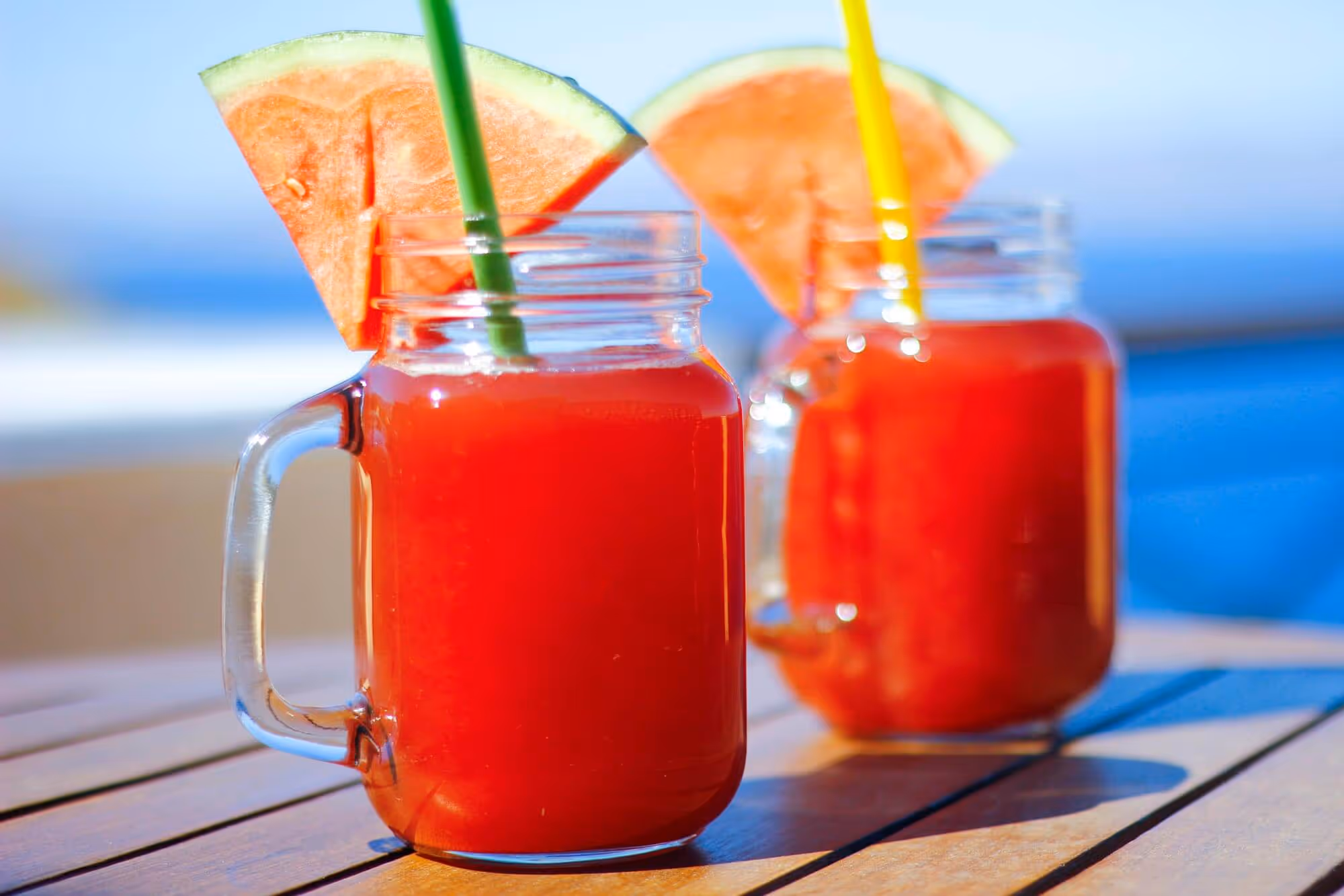 Two glass jars filled with red watermelon juice, each garnished with a watermelon slice and a colored straw, placed on a wooden table.