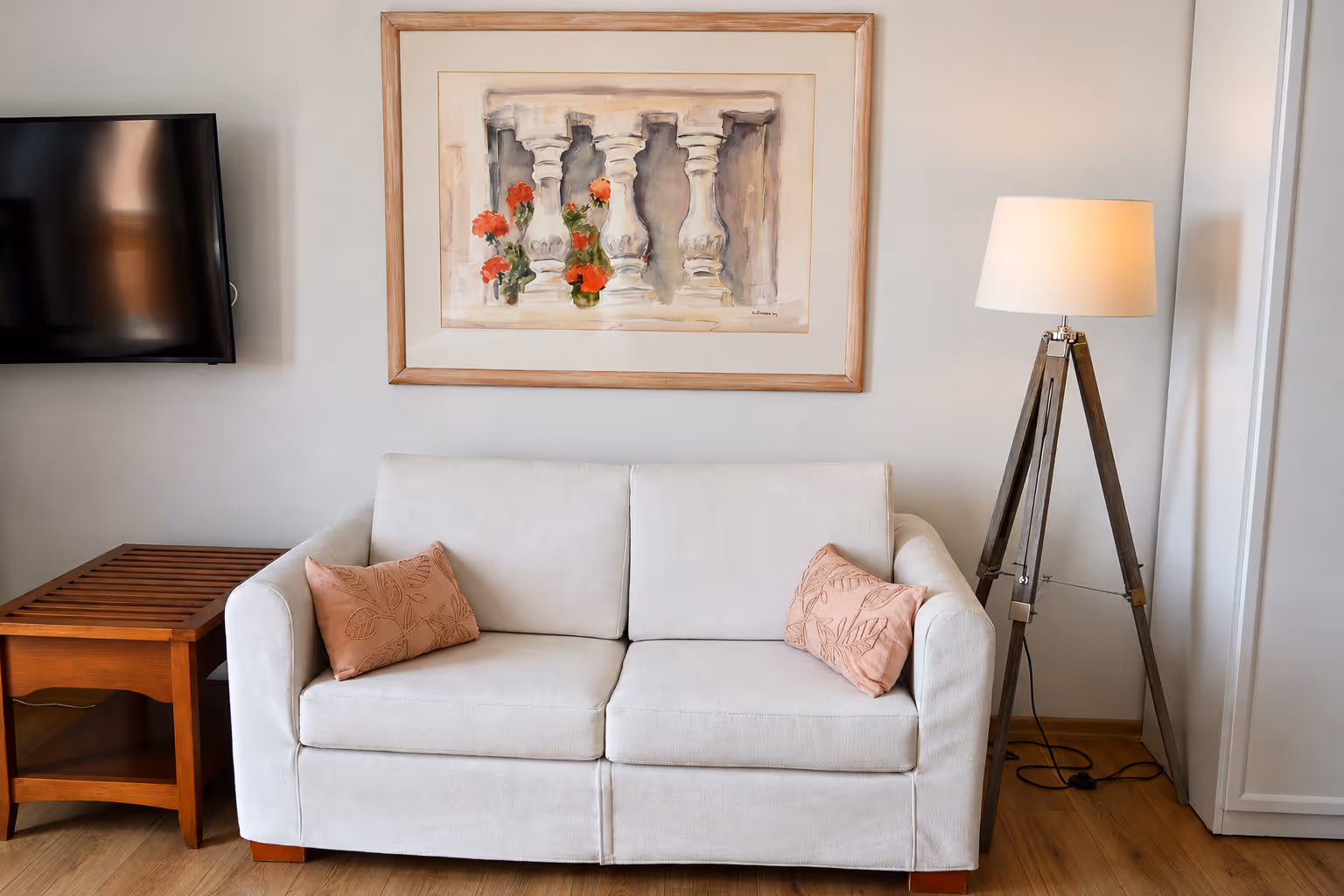 Living room with white loveseat, two pink pillows, wooden side table, floor lamp, and framed artwork.