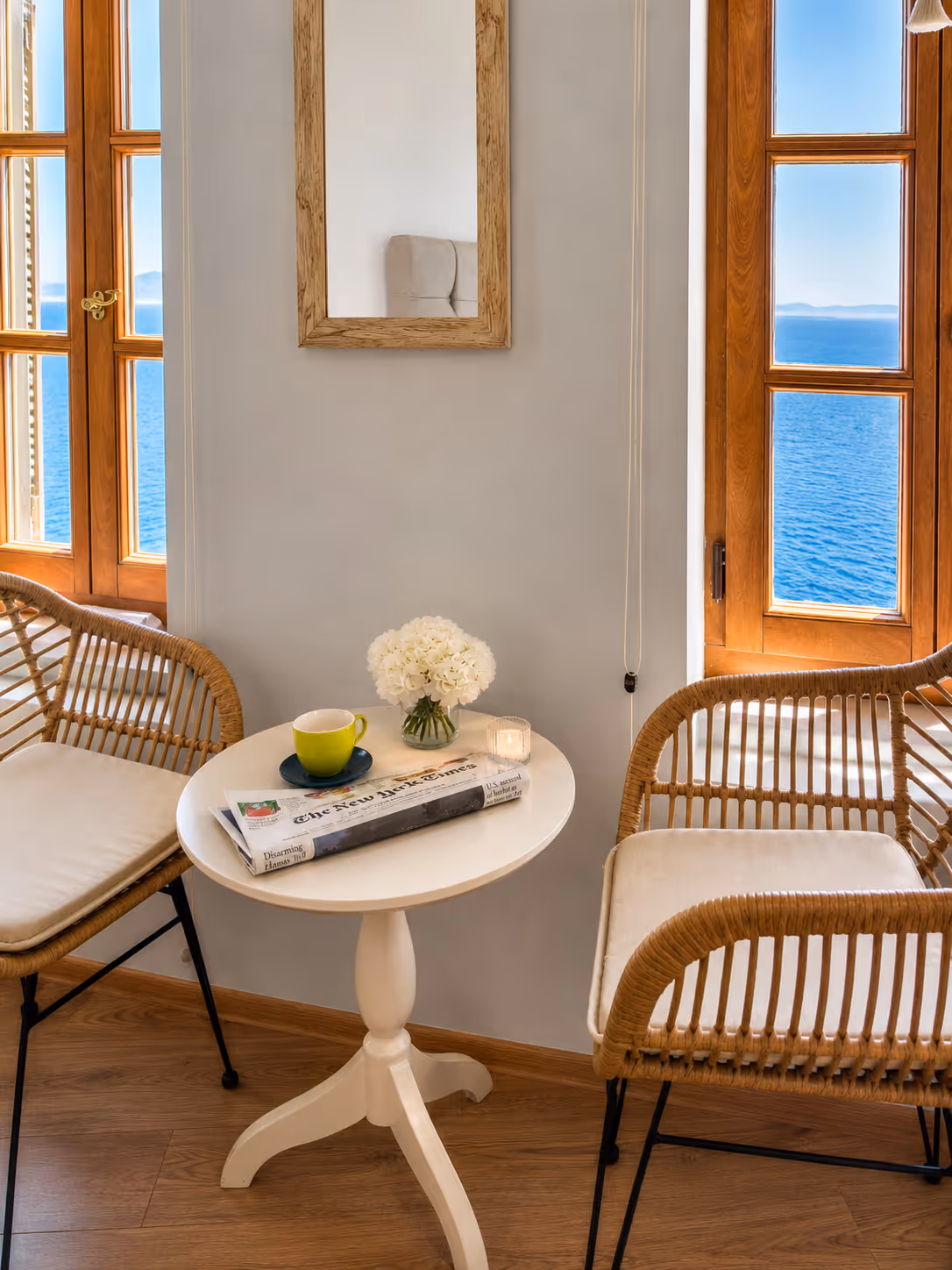 Cozy corner with white round table, two wicker chairs, cup, newspaper, flowers, sea view windows.