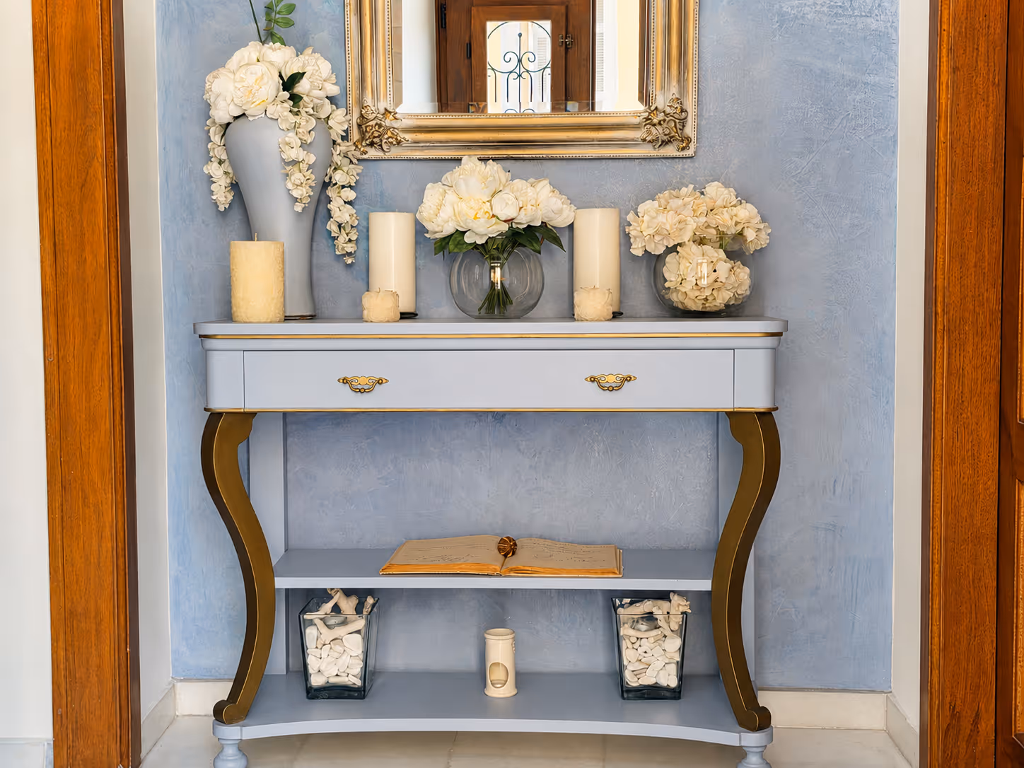 Blue and gold console table with white floral arrangements, candles, and an open book below a gold-framed mirror.