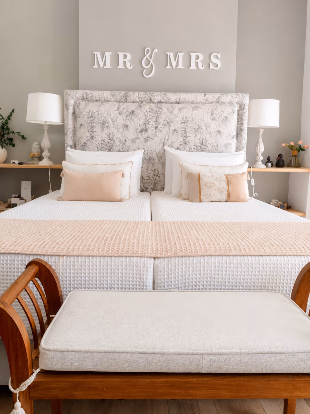 Neutral bedroom with twin beds, floral headboard, 'MR & MRS' wall sign, white lamps, and wooden bench.