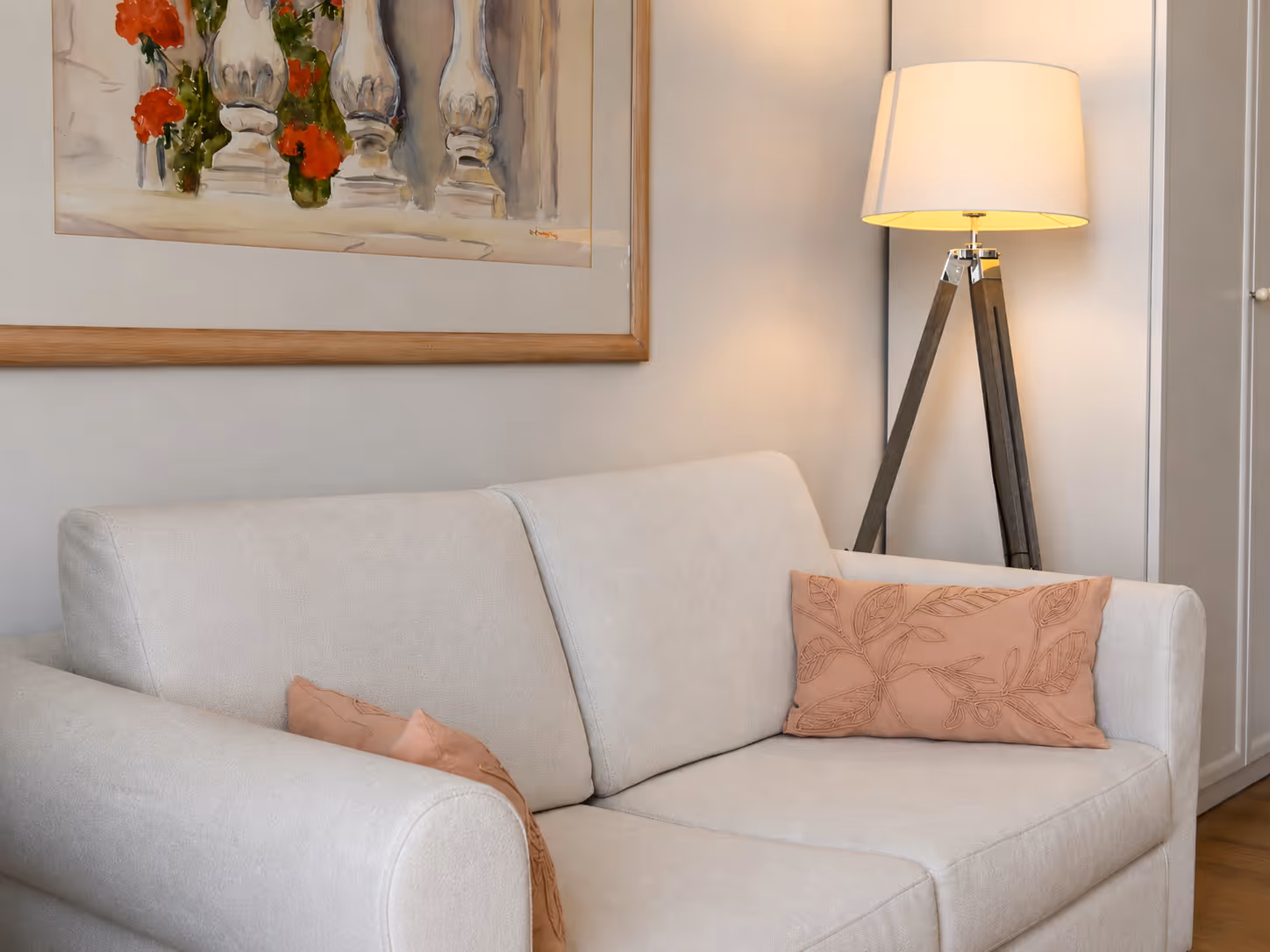 Light beige sofa with two embroidered peach pillows beside a wooden tripod floor lamp and wall art.