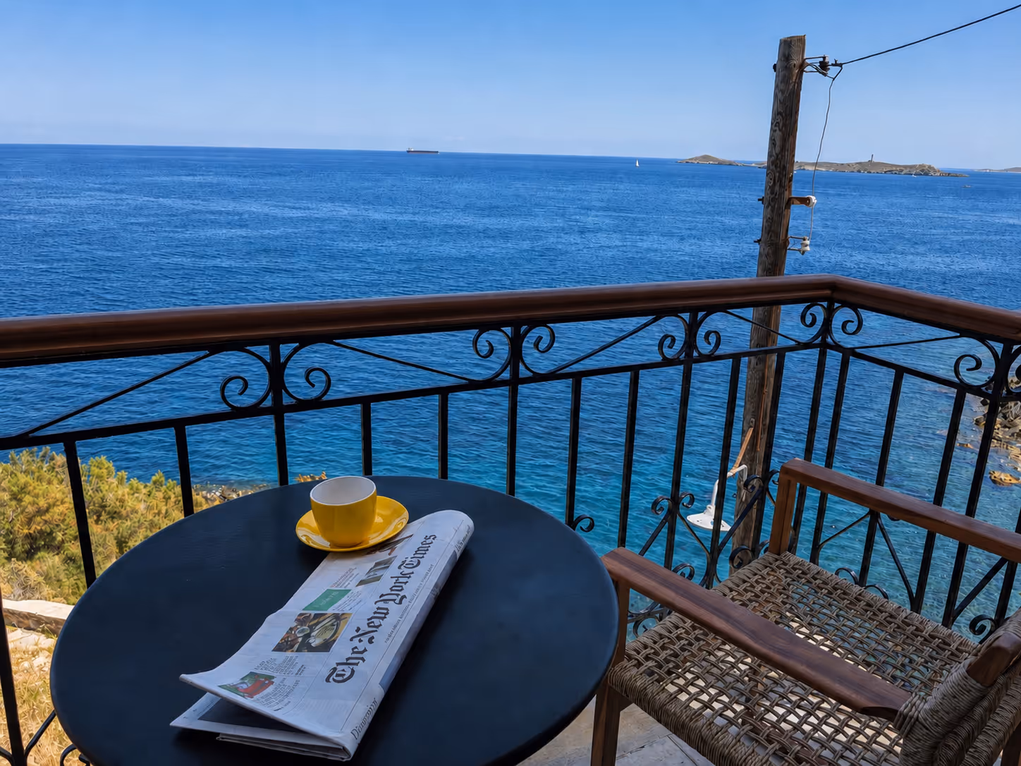 Balcony with a round table holding a yellow cup and The New York Times by the blue sea.