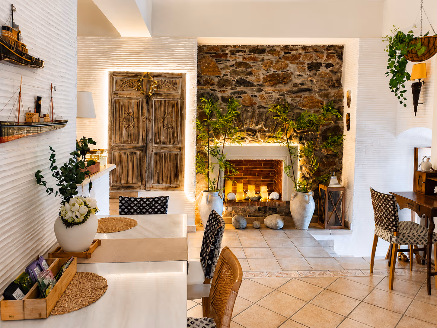 Cozy room with white walls, rustic wooden doors, a stone fireplace, plants, and dining tables with chairs.