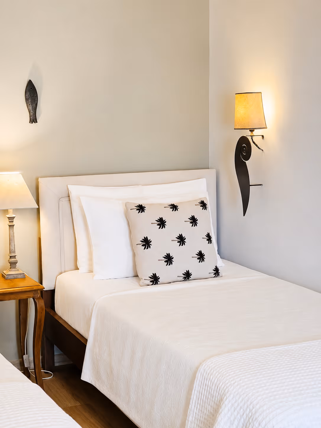 White bed with palm tree patterned pillow, bedside table with lamp, and wall sconce in bedroom.