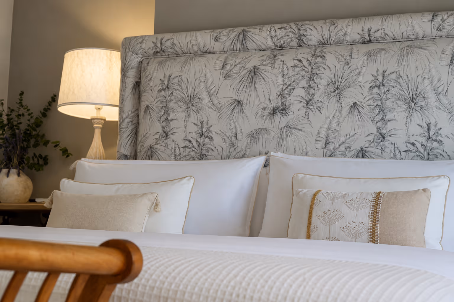 Bed with white pillows, patterned cushions, floral upholstery headboard, and a lit lamp on a side table.