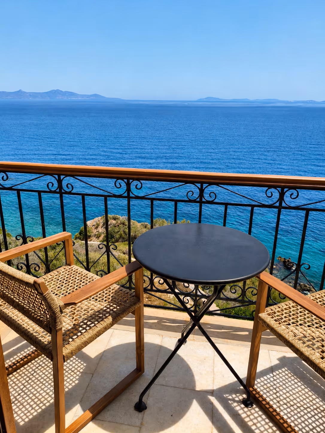 Two woven wooden chairs and a small black table on a balcony overlooking blue ocean and distant mountains.