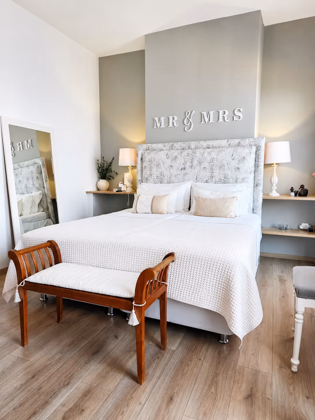 Bright bedroom with a large bed, wooden bench, floral headboard, and MR & MRS sign on wall.