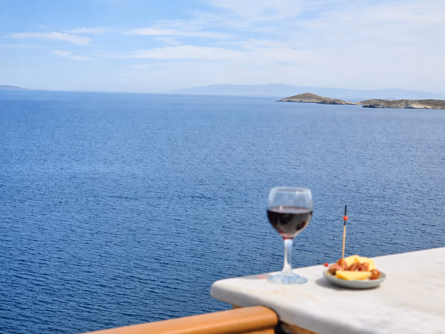 Glass of red wine and a snack plate on a balcony overlooking calm blue sea and distant islands.