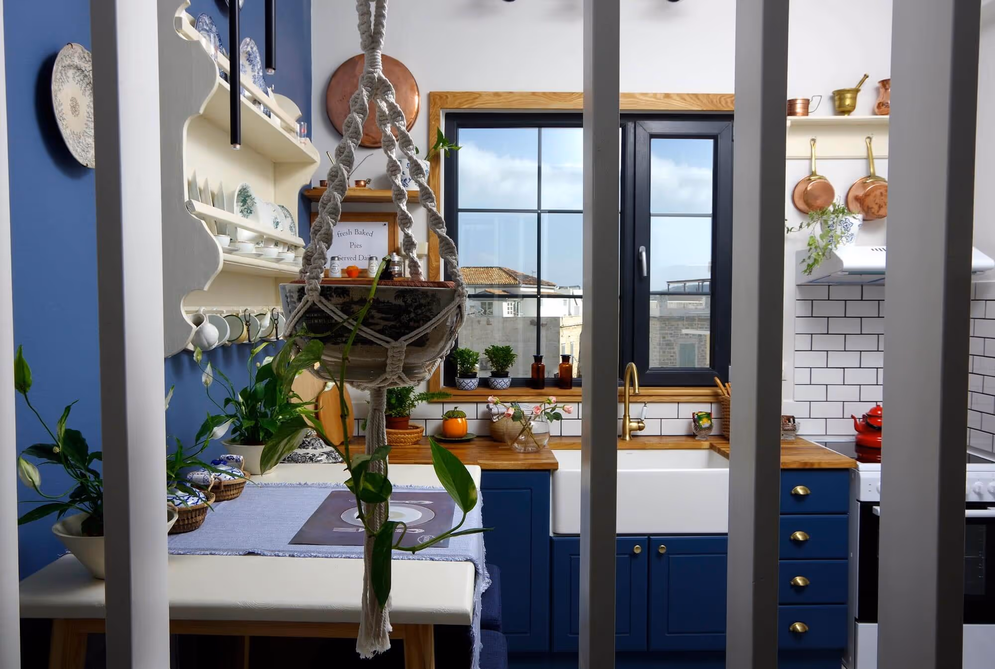 Cozy kitchen with blue cabinets, white farmhouse sink, wooden countertops, hanging plant, and window overlooking buildings.