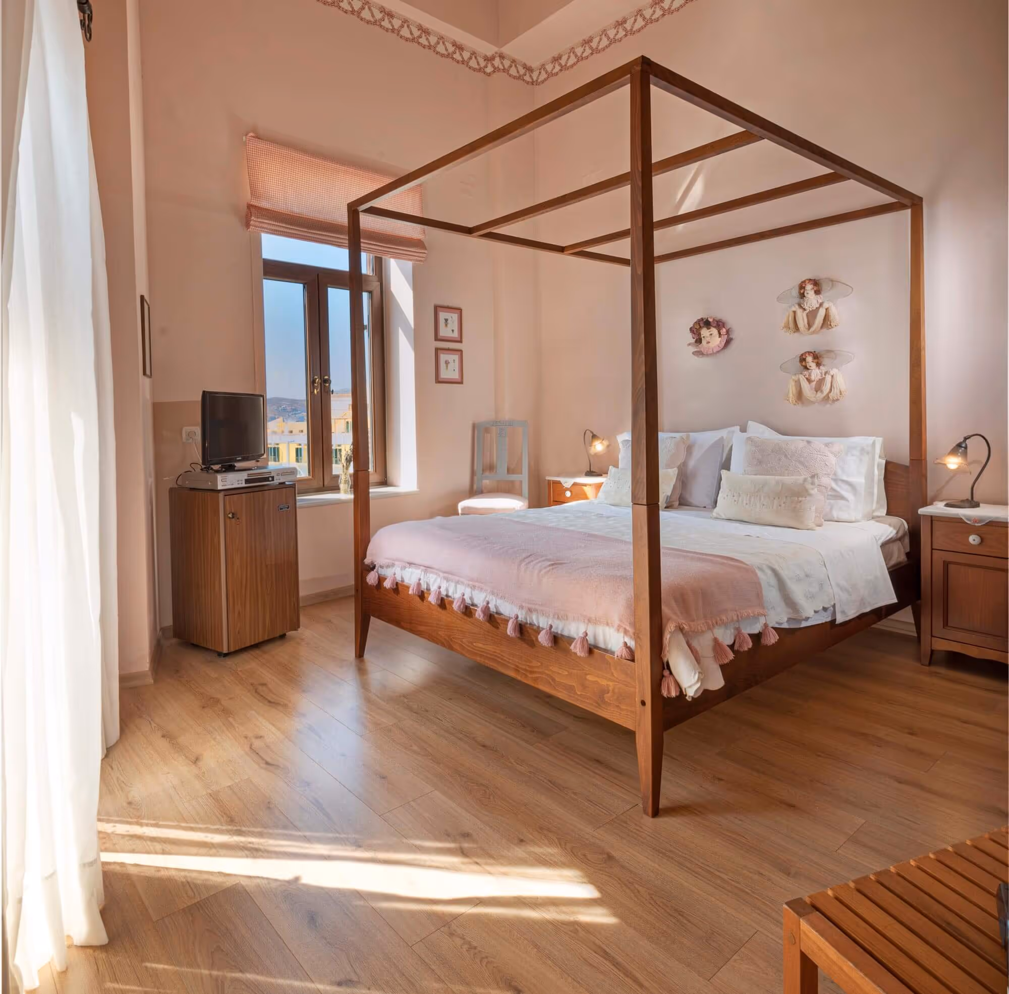 Bedroom with a wooden canopy bed dressed with white and pink bedding, two wooden nightstands with lamps, a chair by the window, and wall decorations of three angel figures.