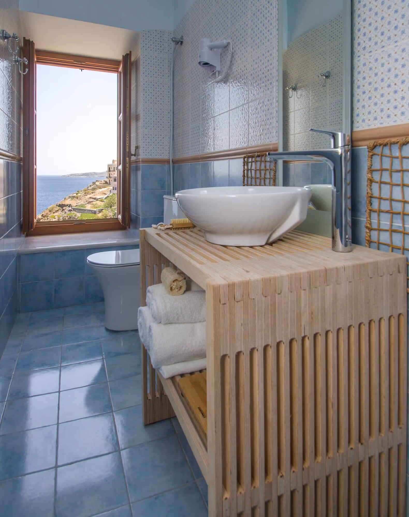 Small bathroom with blue tile floor and walls, wooden vanity with white vessel sink, towels on shelf, and open window showing sea view.